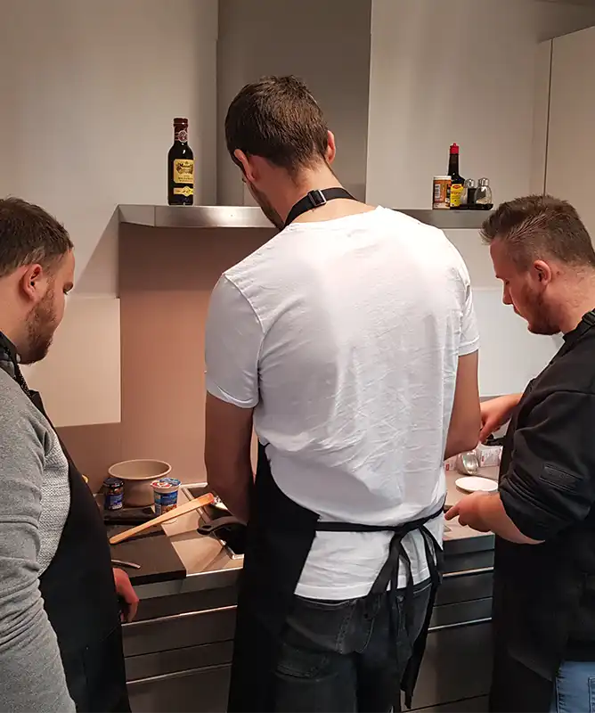 Drei Männer in Egenberger-Schürzen kochen gemeinsam an einem Küchenherd mit Zutaten aus der Dose.