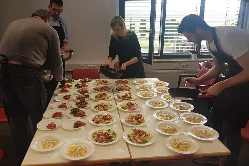 Vier Personen, darunter Egenberger, stellen in einem hellen Raum verschiedene Gerichte auf einen Tisch.