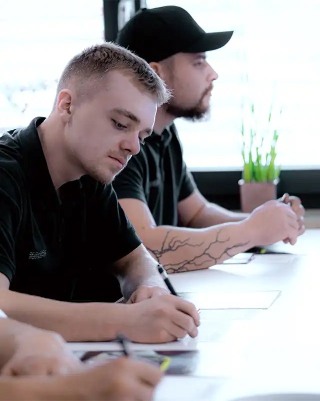 Zwei Männer in dunklen Hemden schreiben an einem Tisch mit einem Egenberger-Becher und einer Topfpflanze.