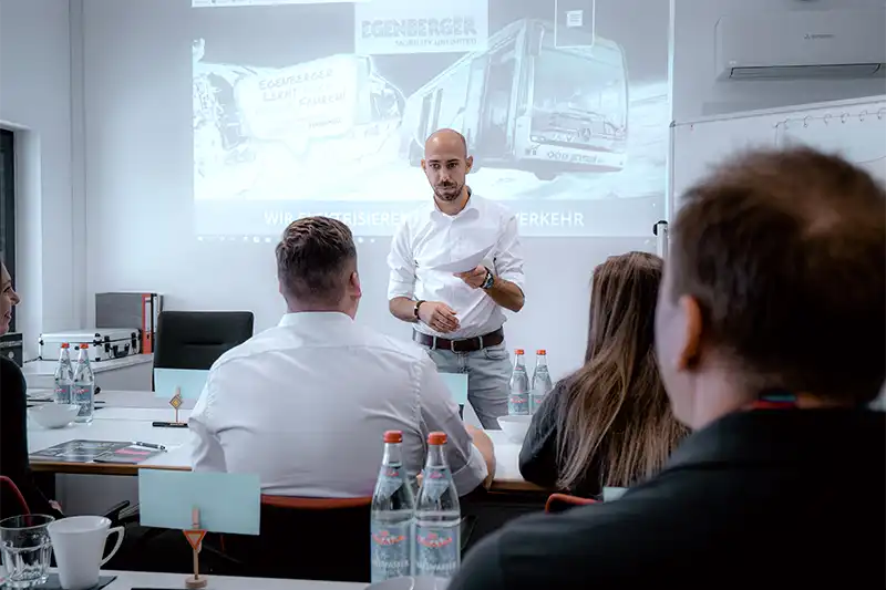 Ein Mann, möglicherweise Egenberger, spricht zu einer sitzenden Gruppe, hinter der eine Präsentation gezeigt wird.