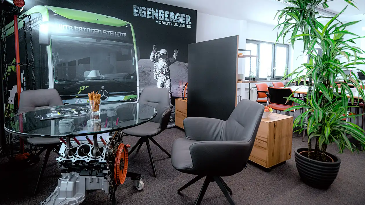 Modernes Büro: Glastisch auf Egenberger-Motorteilen, graue Stühle, Pflanze, Bus/Astronauten-Wandbild.